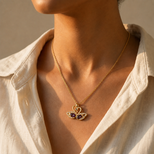 A close-up of a model wearing a gold toned amora teddy demi fine chain necklace with a swan pendant featuring purple gemstone wings, styled with an off-white linen shirt.