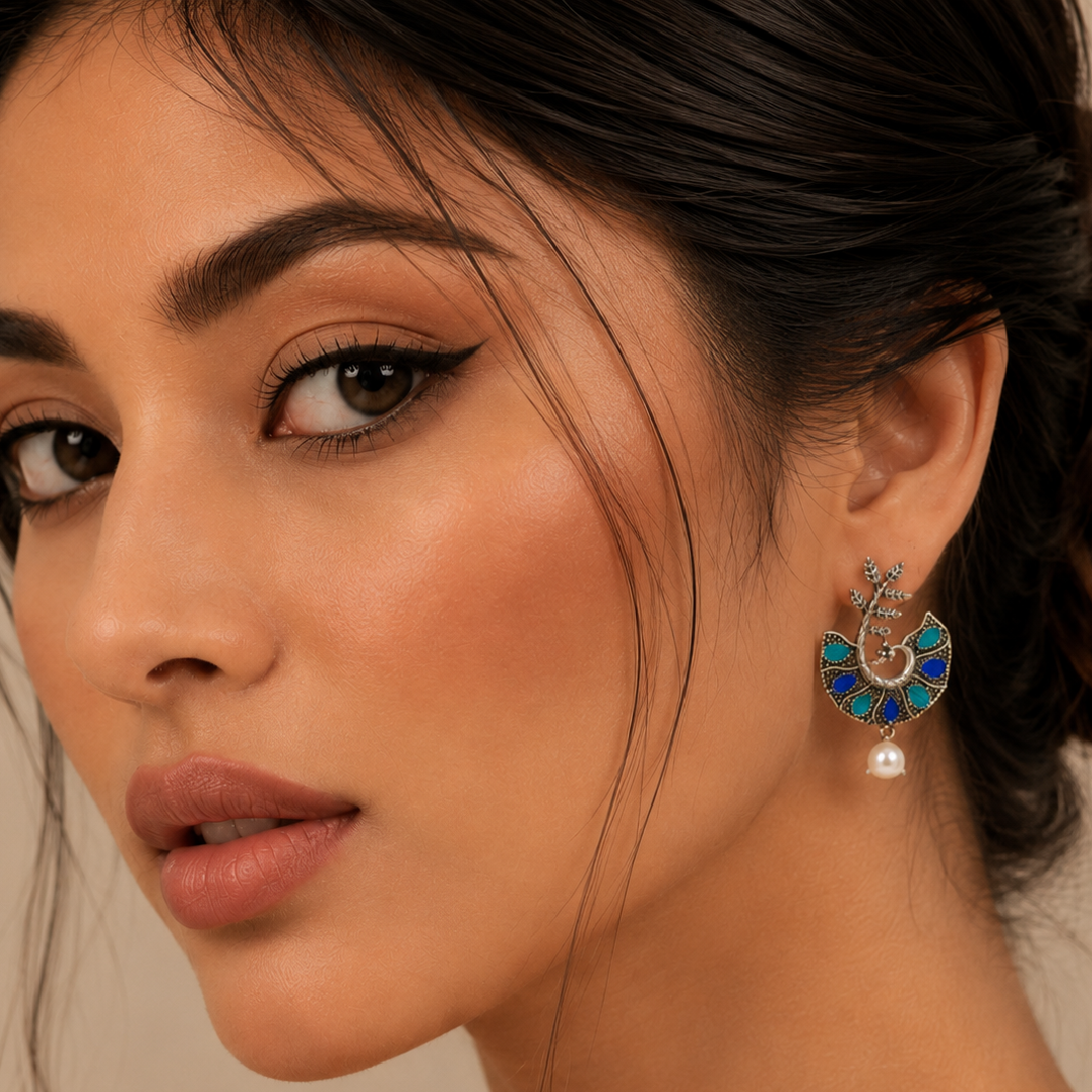 A close-up of a woman wearing a dangling statement earring. The earring features a silver-toned crescent moon and floral design embellished with blue and green enamel, with a small white pearl drop at the bottom.