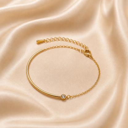 A gold-toned bangle bracelet with a single clear gemstone, displayed on a draped champagne-colored satin fabric.
