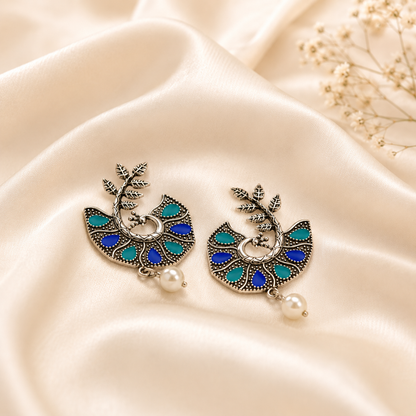 A pair of bohemian-style dangle earrings with blue and teal enamel details, silver-toned metalwork, and a dangling pearl, displayed on a cream satin fabric background.