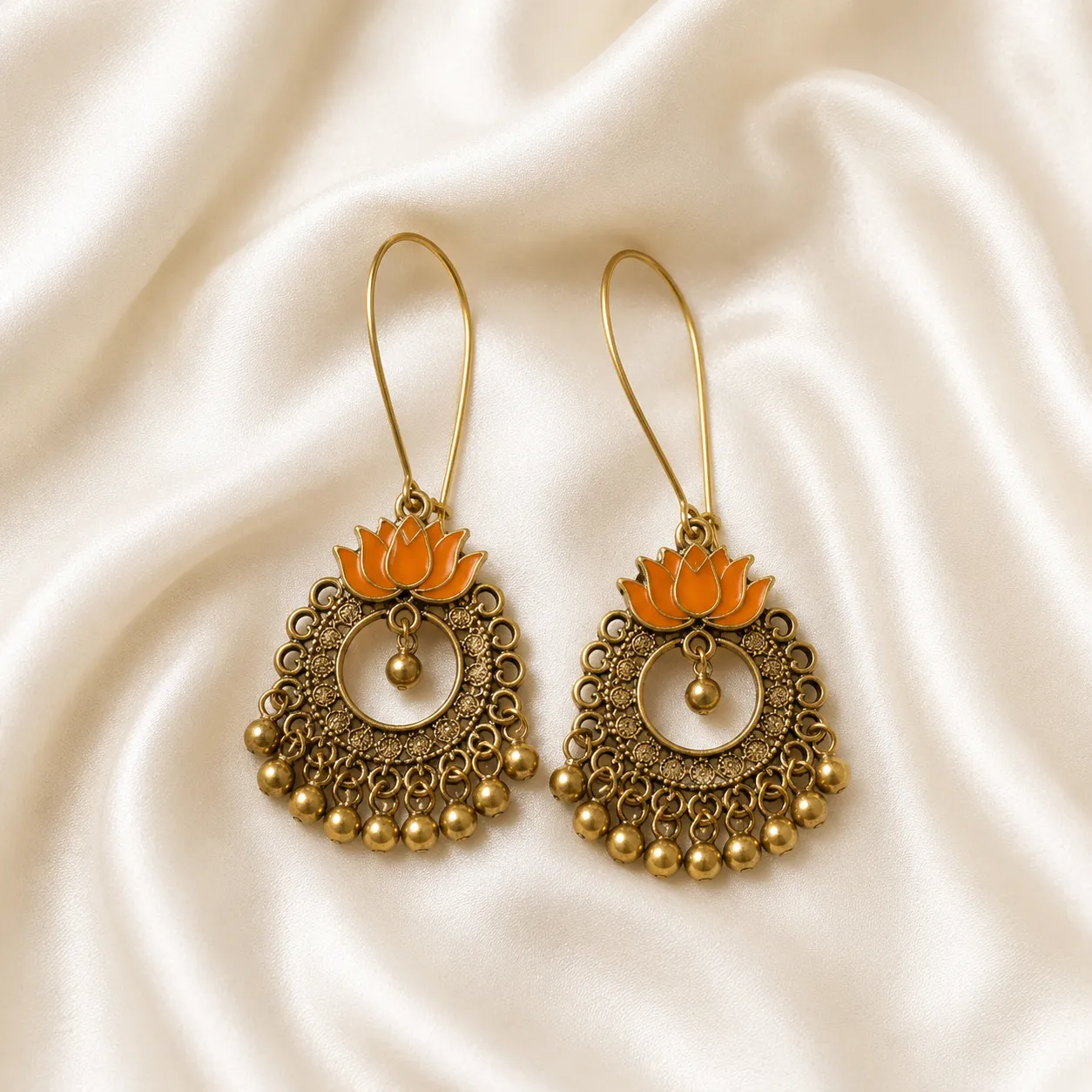 A pair of bohemian-style gold-toned dangle earrings with orange lotus flower charms, displayed on a draped cream satin fabric background.