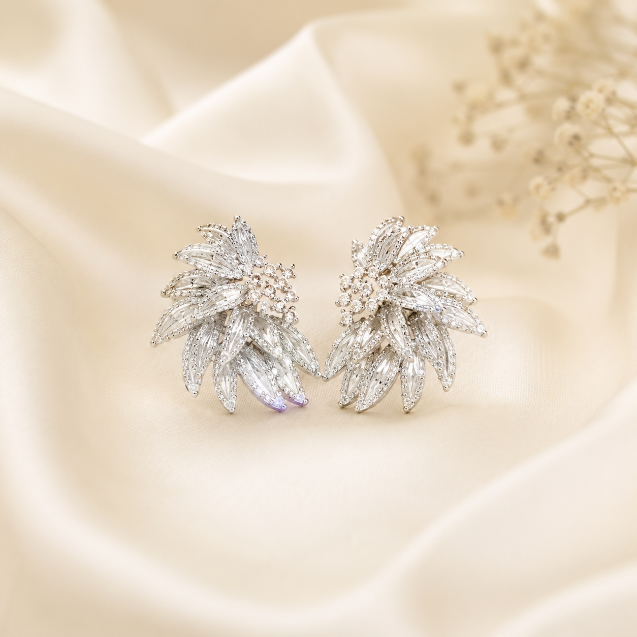 A pair of silver-toned leaf-shaped stud earrings adorned with clear pavé stones, displayed on draped cream satin fabric with small dried flowers in the background.