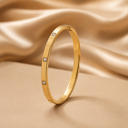 A slim gold tonned finish bangle bracelet with three small square-cut clear stones spaced along the band, photographed on a warm beige surface with draped satin fabric in the background.