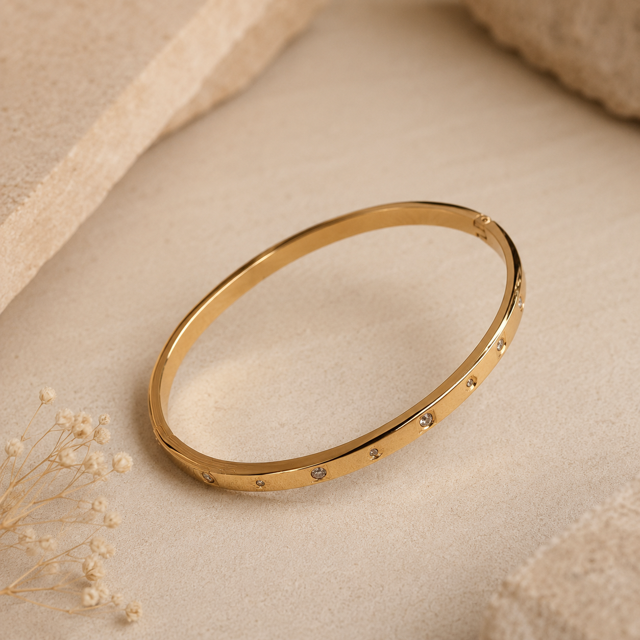 A thin gold tonned bangle bracelet with small inset stones, displayed on a textured beige fabric surface with dried flowers and soft neutral props.