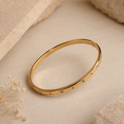 A thin gold tonned bangle bracelet with small inset stones, displayed on a textured beige fabric surface with dried flowers and soft neutral props.