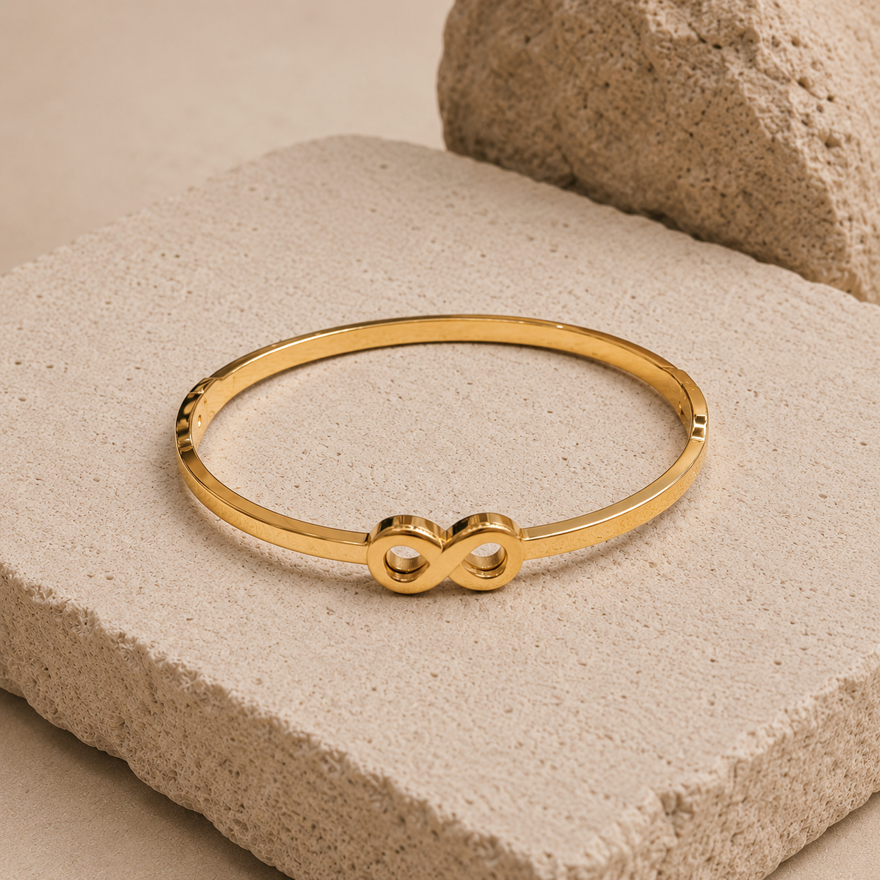 A thin gold tonned finish bangle bracelet with an infinity symbol at the center, displayed on a textured beige stone block.