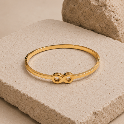 A thin gold tonned finish bangle bracelet with an infinity symbol at the center, displayed on a textured beige stone block.