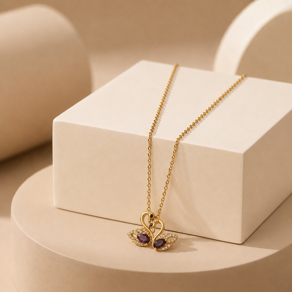 A gold-tone amora teddy anti tarnish necklace with a butterfly pendant featuring purple gemstones and small clear stones, displayed on a white geometric block against a warm beige studio background.