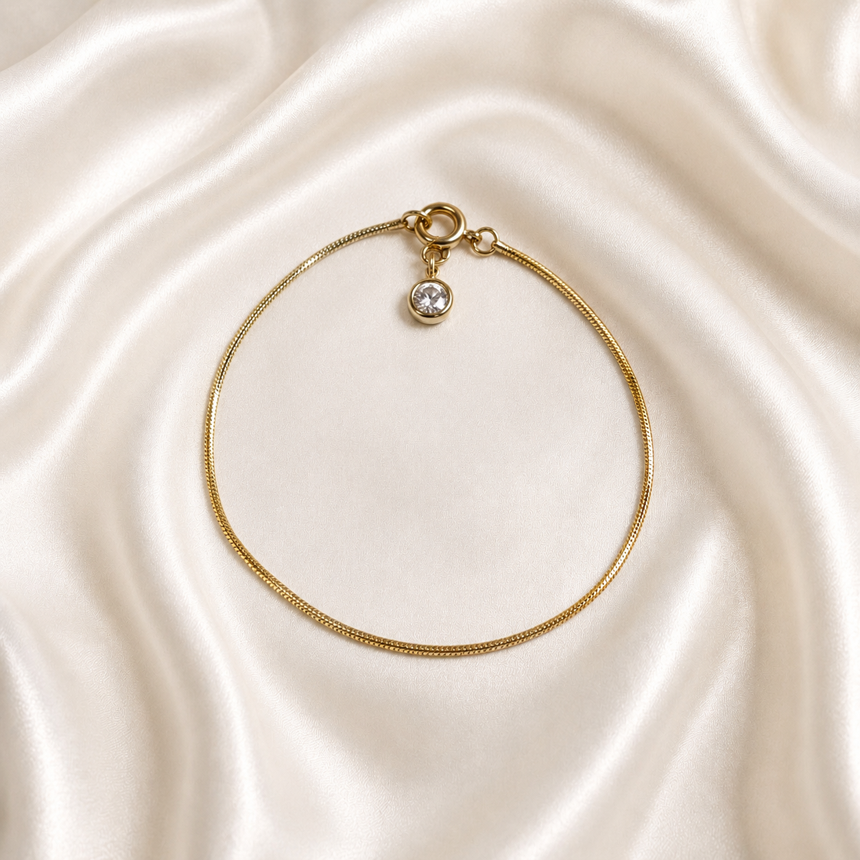 A delicate gold tonned Aura Lock Bracelet with a small round clear gemstone charm, displayed on a draped cream satin fabric background.