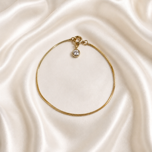 A delicate gold tonned Aura Lock Bracelet with a small round clear gemstone charm, displayed on a draped cream satin fabric background.