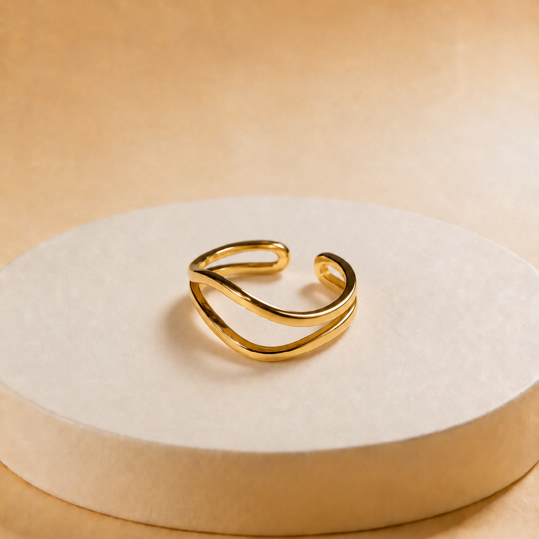 A gold-toned open cuff ring with a smooth, flowing double-band design, displayed on a round beige pedestal against a warm beige background.
