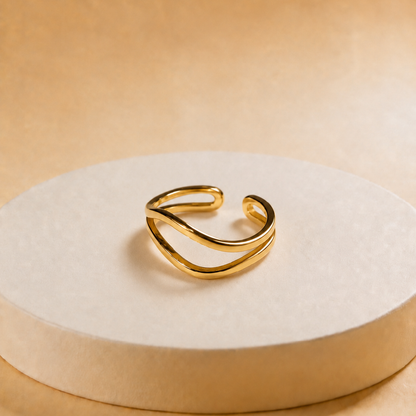 A gold-toned open cuff ring with a smooth, flowing double-band design, displayed on a round beige pedestal against a warm beige background.