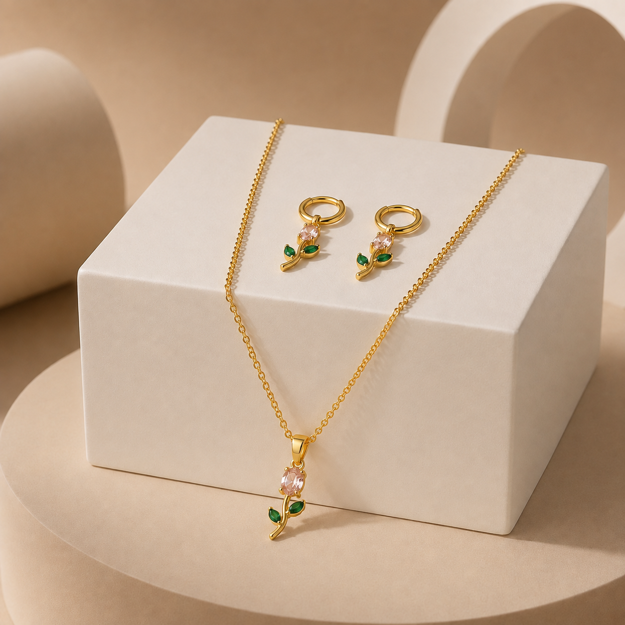 Gold-tone necklace and matching drop earrings with pale pink stones and green leaf accents, displayed on a white rectangular block against a warm beige studio background.