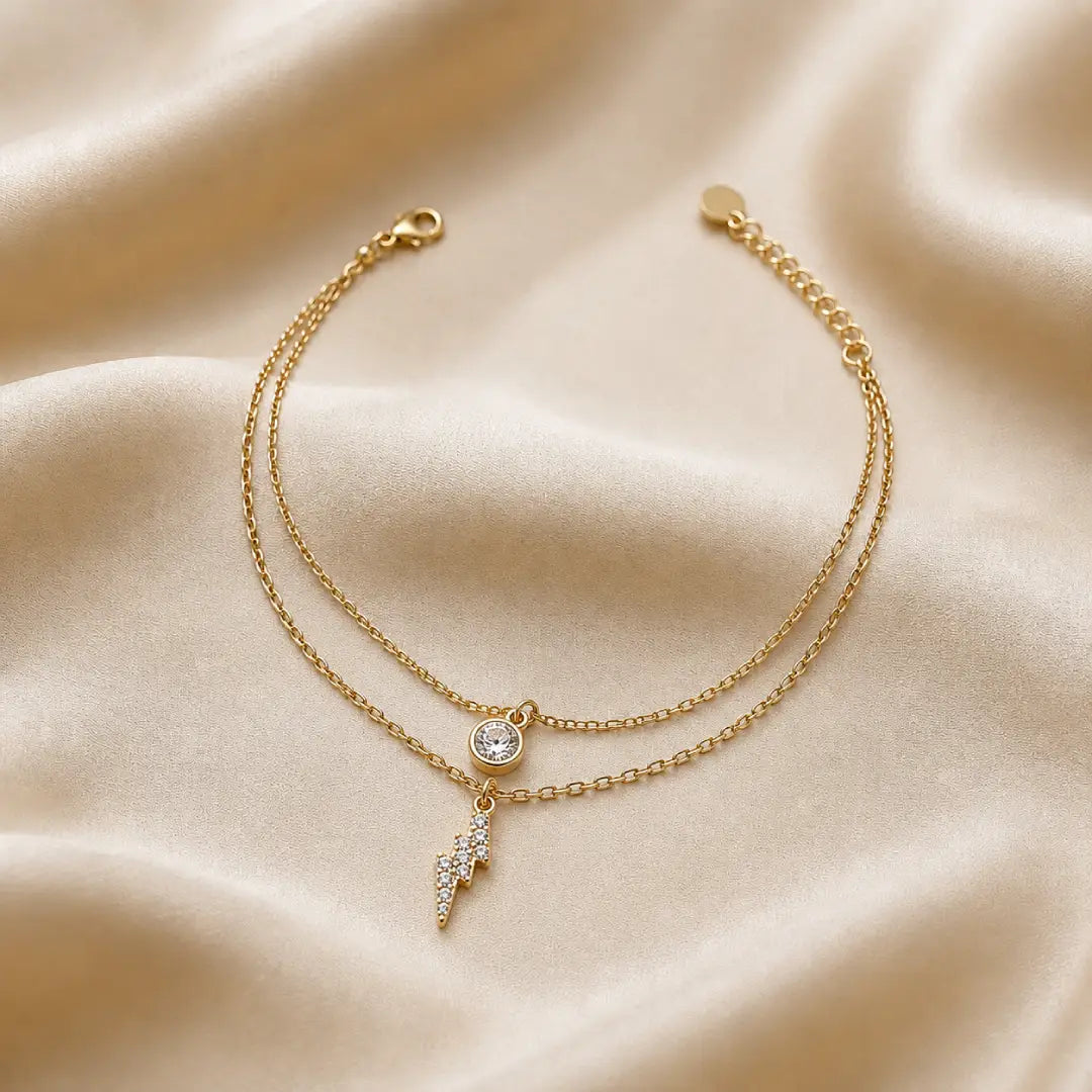 Gold tonned aura dual layer bracelet with a small round charm and lightning bolt charm on a beige fabric background