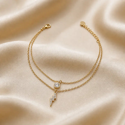 Gold tonned aura dual layer bracelet with a small round charm and lightning bolt charm on a beige fabric background