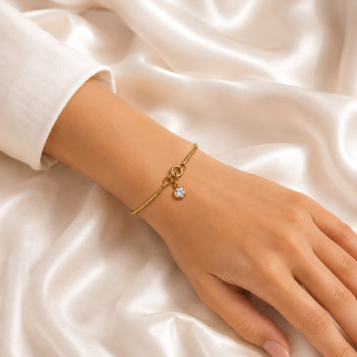 Gold tonned anti tarnish aura lock bracelet with a small clear crystal charm, worn on a person's wrist against a draped cream satin fabric background.