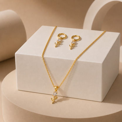Gold tonned jewelry set featuring a delicate chain necklace and matching hoop earrings, both adorned with small clear crystal-like accents and leaf-shaped pendants, displayed on a white rectangular pedestal against a warm beige studio background.