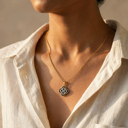 A close-up of a person wearing a delicate gold chain necklace with a black and white clover pendant, styled with an open white linen shirt.