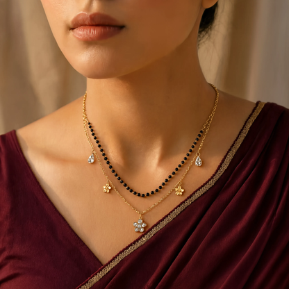 A close-up of a woman wearing layered gold necklaces, including a black beaded necklace, a delicate gold chain with small floral charms, and a pendant necklace, paired with a deep maroon velvet top.