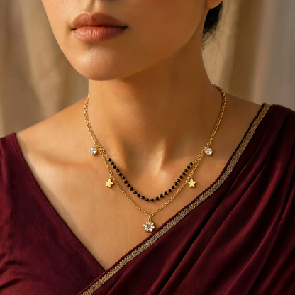 A close-up of a woman wearing a layered gold tonned necklace with black beads, small star charms, and a flower-shaped pendant, paired with a deep maroon draped garment.