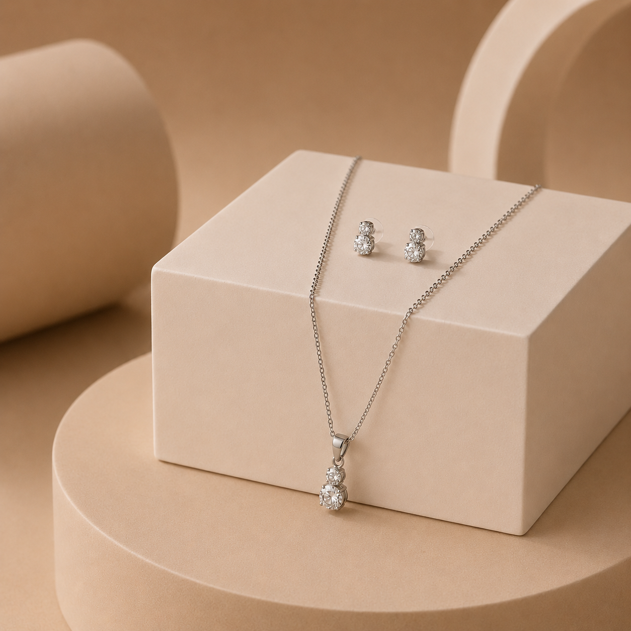 Silver-toned jewelry set featuring a delicate chain necklace with a pear-cut gemstone pendant and matching stud earrings, displayed on a beige geometric pedestal.