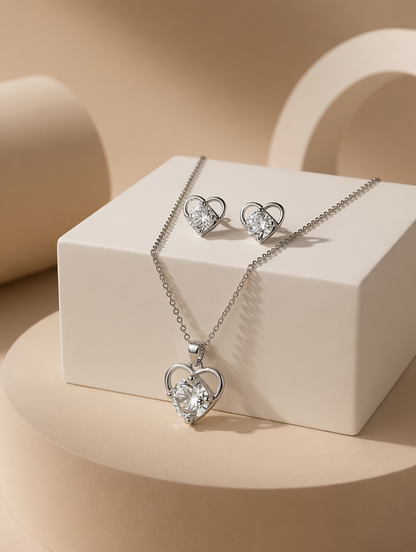 Silver tonned heart-shaped pendant necklace and matching stud earrings with clear heart-shaped gemstones, displayed on a white geometric pedestal against a warm beige background.