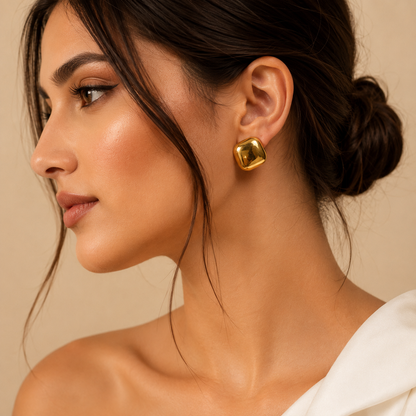 A close-up side profile of a woman wearing a single shiny gold square stud earring with softly rounded corners, set against a warm beige background.