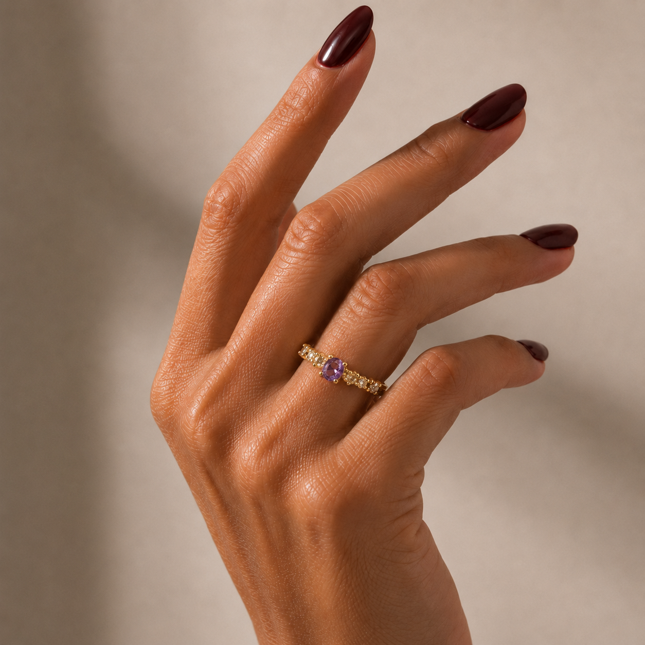 A close-up of a hand with dark burgundy manicured nails wearing a gold-tonned ring with a central purple gemstone and small clear stones on either side, set against a warm beige background.