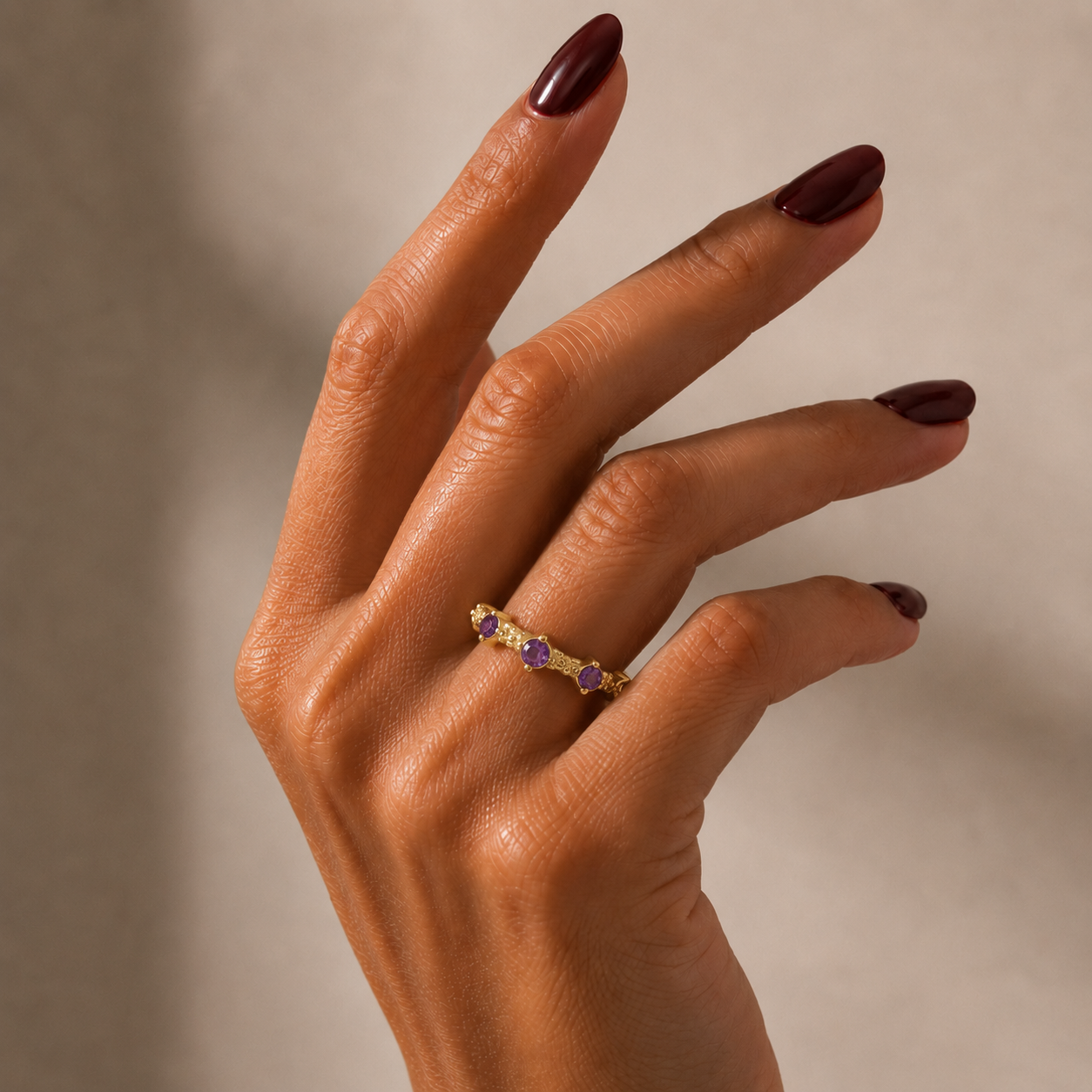 A close-up of a hand with dark burgundy nail polish wearing a delicate gold-tonned ring set with small yellow and purple gemstones, set against a soft beige background.