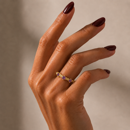 A close-up of a hand with dark burgundy nail polish wearing a delicate gold-tonned ring set with small yellow and purple gemstones, set against a soft beige background.