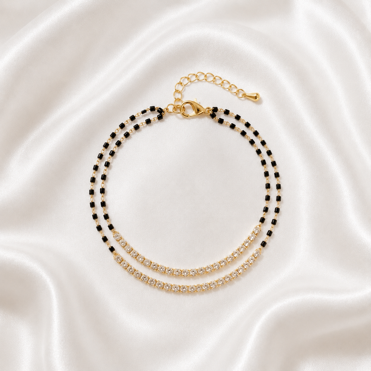 A double-strand beaded anklet with black and gold beads, featuring a gold-tone lobster clasp and extender chain, displayed on a draped white satin fabric background.