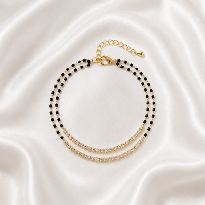 A double-strand beaded anklet with black and gold beads, featuring a gold-tone lobster clasp and extender chain, displayed on a draped white satin fabric background.