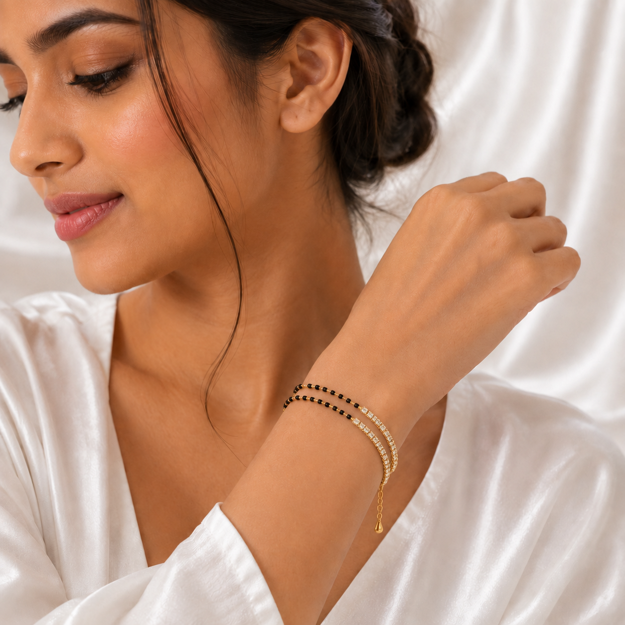 A woman wearing a delicate double-strand bracelet with black and white beads and a small gold charm, shown in a close-up lifestyle shot against a white satin background.