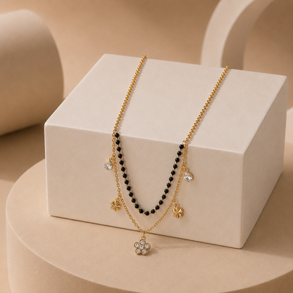 A double-strand gold tonned necklace displayed on a cream-colored jewelry box. One strand features small black beads, while the other has gold flower charms and a central flower pendant embellished with clear rhinestones.