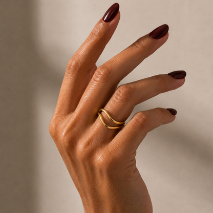 A close-up of a hand with dark burgundy nail polish wearing a thin, double-band open cuff ring against a neutral beige background.