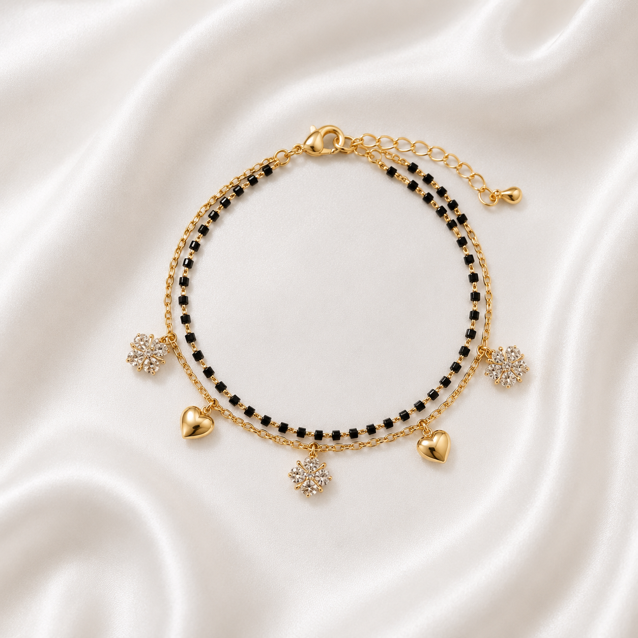 A gold-tone double-strand beaded bracelet with black beads, heart charms, and floral crystal charms, displayed on a white satin fabric background.