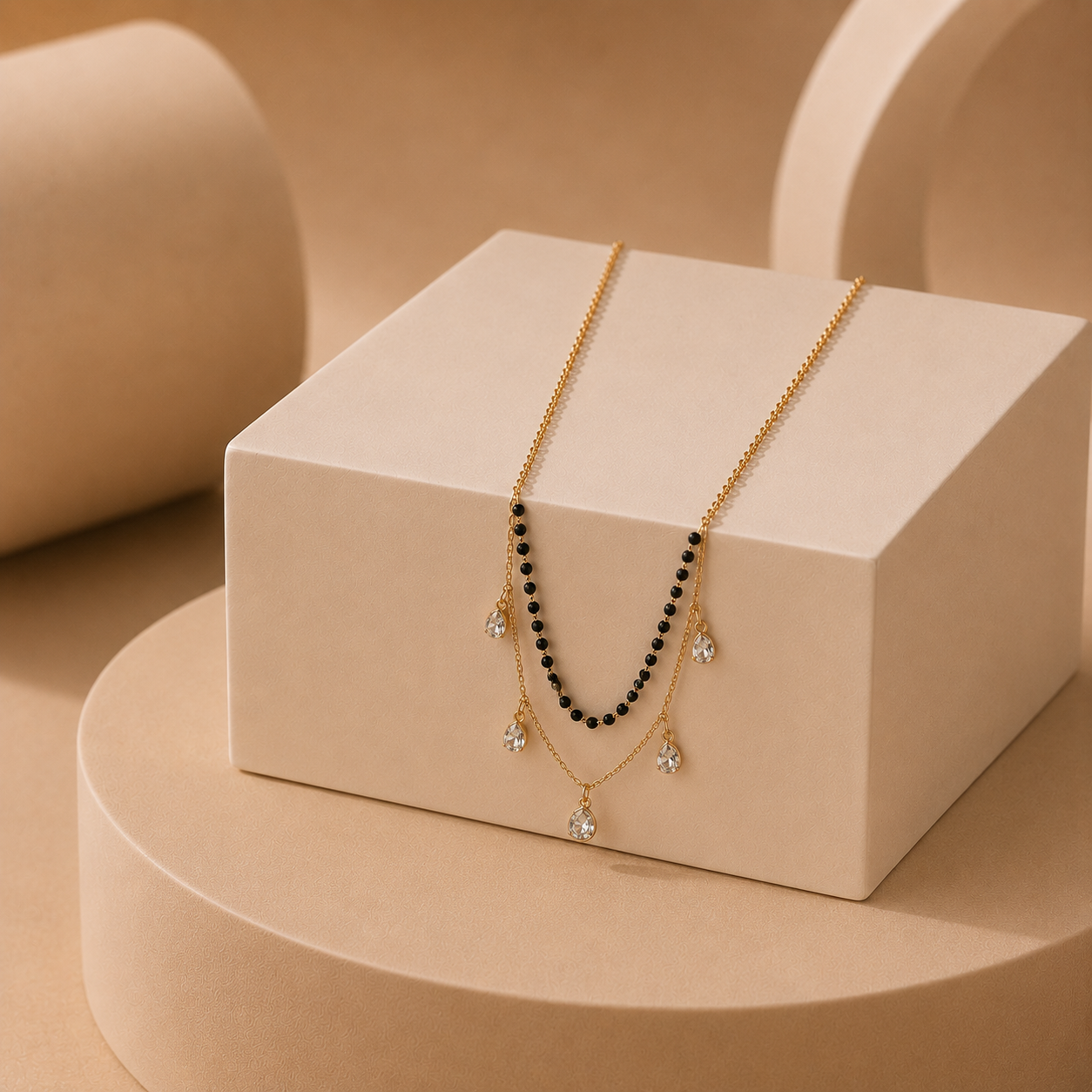 A gold-tone layered necklace with a black beaded strand and clear teardrop gemstones, displayed on a cream-colored block against a warm beige background.