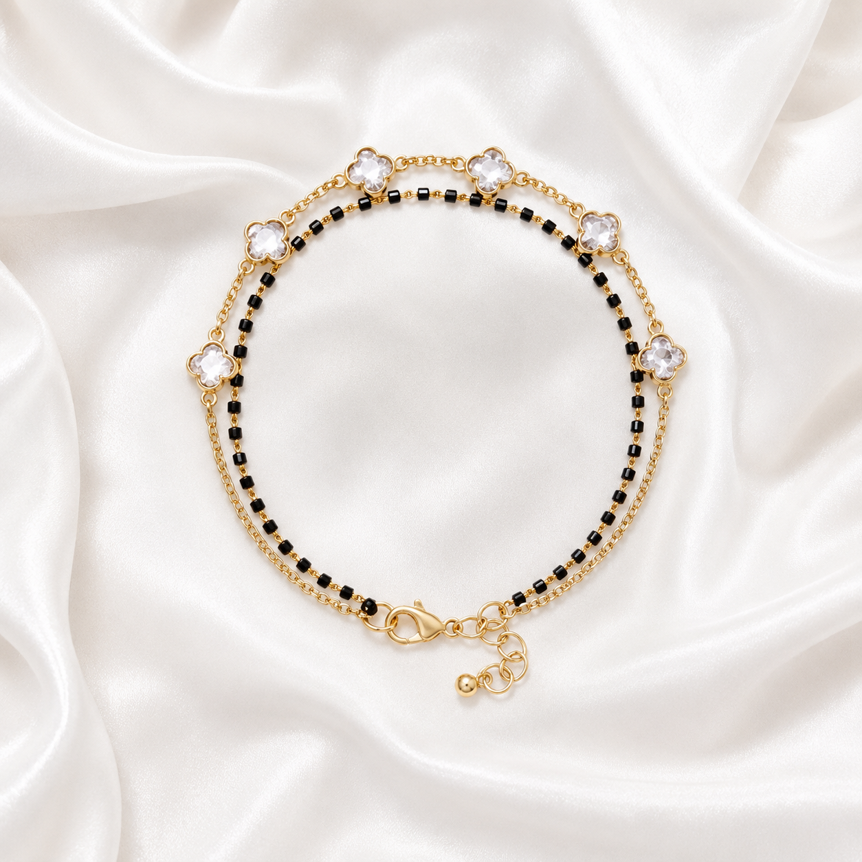 A gold-toned beaded bracelet with alternating rows of small black beads and larger clear rhinestone flower motifs, displayed on a draped white satin fabric background.