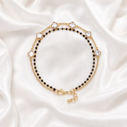 A gold-toned beaded bracelet with alternating rows of small black beads and larger clear rhinestone flower motifs, displayed on a draped white satin fabric background.