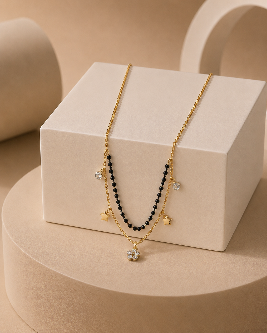 A gold-toned double-strand necklace draped over a white display block. One strand features small black beads, while the other has gold star charms and a round clear crystal pendant.