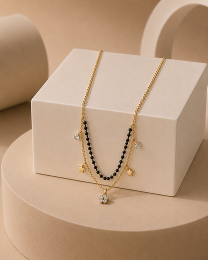 A gold-toned double-strand necklace draped over a white display block. One strand features small black beads, while the other has gold star charms and a round clear crystal pendant.