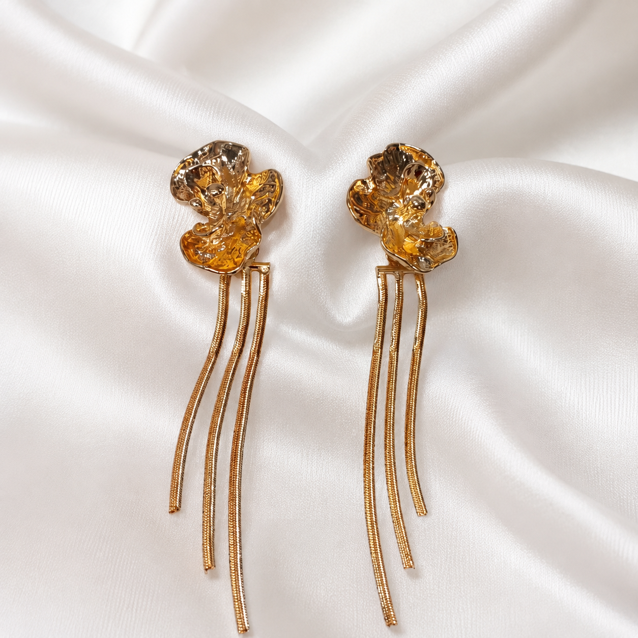A pair of gold-toned flower-shaped earrings with faceted amber-yellow crystal tops and three dangling curved wire strands, displayed on draped white satin fabric.