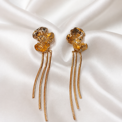 A pair of gold-toned flower-shaped earrings with faceted amber-yellow crystal tops and three dangling curved wire strands, displayed on draped white satin fabric.