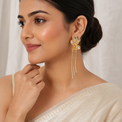 A woman wearing a gold-toned traditional Indian jhumka earring with dangling chains, shown in a close-up portrait against a soft white curtain background.