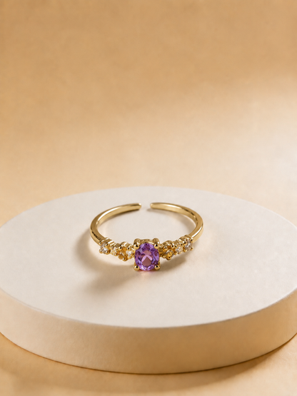 A gold-tonned adjustable ring with a central purple oval gemstone and small clear accent stones on either side, displayed on a round beige pedestal against a warm beige background.