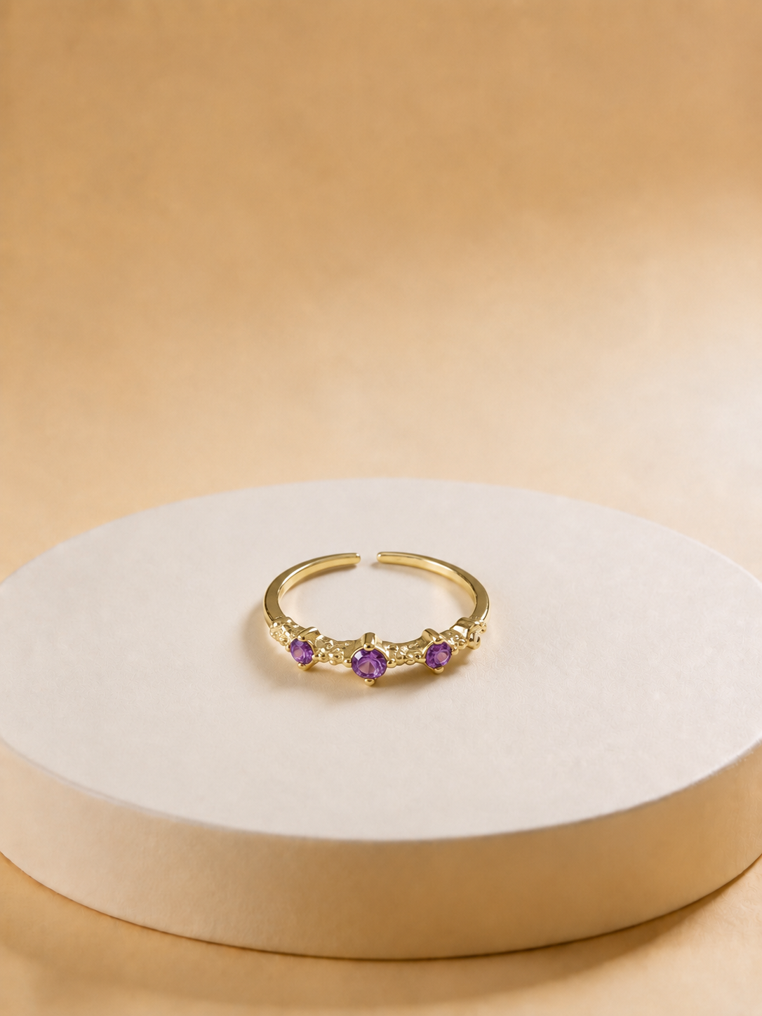 A gold-tonned adjustable ring with three small purple gemstones, displayed on a round beige pedestal against a warm beige background.