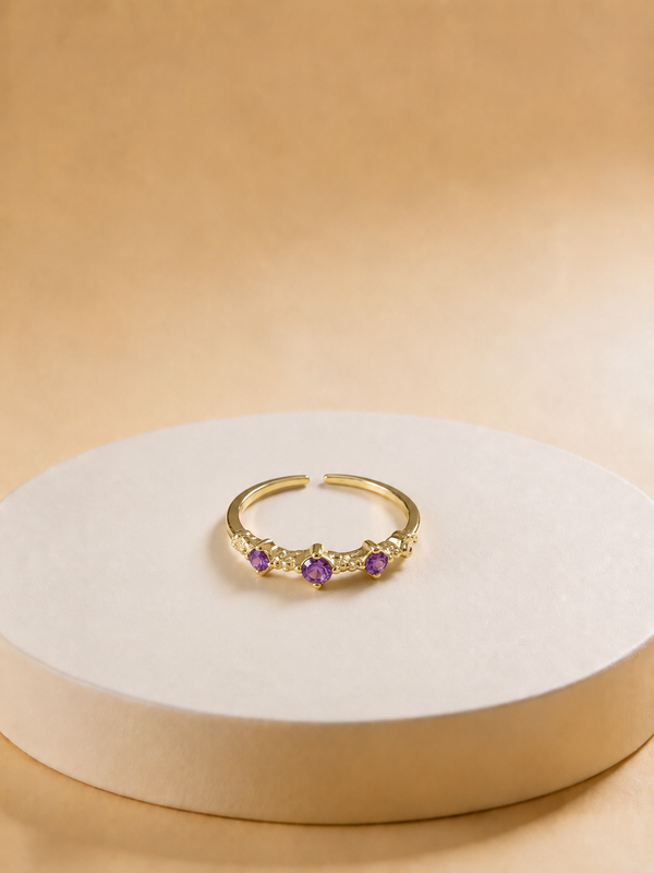 A gold-tonned adjustable ring with three small purple gemstones, displayed on a round beige pedestal against a warm beige background.
