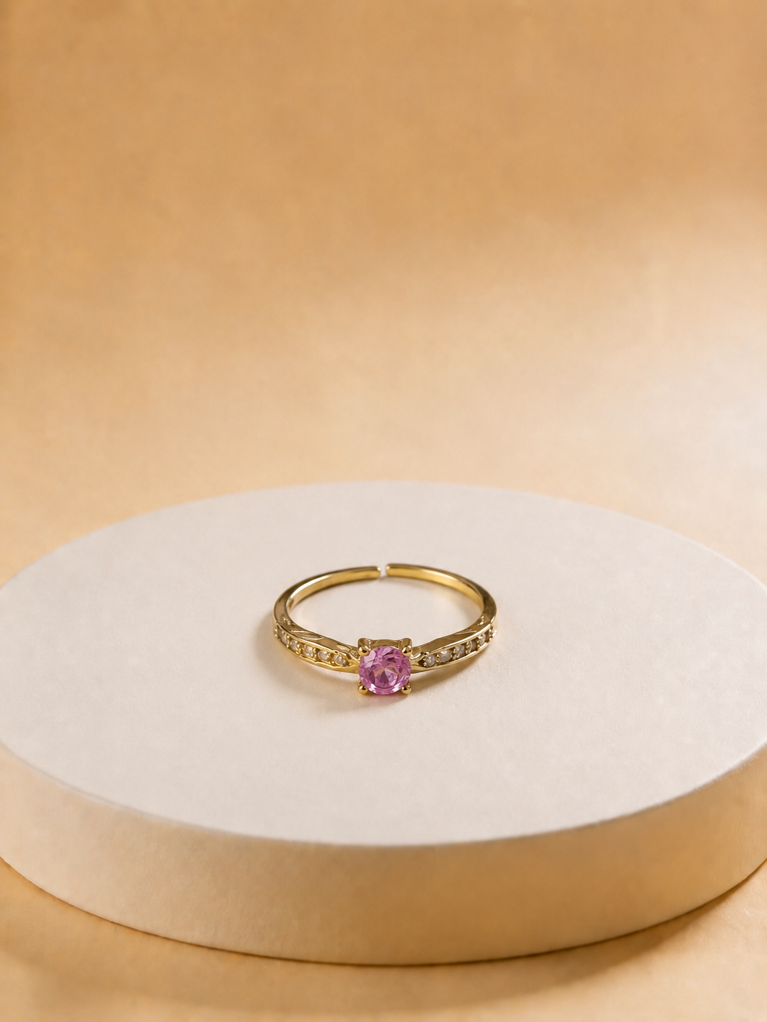 A gold-tonned ring with a central pink gemstone and small side stones, displayed on a round beige pedestal against a warm beige background.