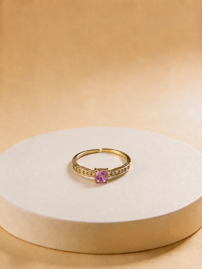 A gold-tonned ring with a central pink gemstone and small side stones, displayed on a round beige pedestal against a warm beige background.