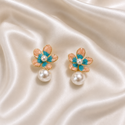 A pair of gold flower-shaped earrings with peach and turquoise enamel petals, a white pearl center, and a dangling white pearl, displayed on draped cream satin fabric.